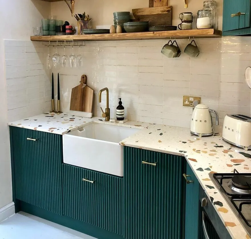 Green fluted modern kitchen cabinets with terrazzo countertop and wooden shelves holding kitchen items.