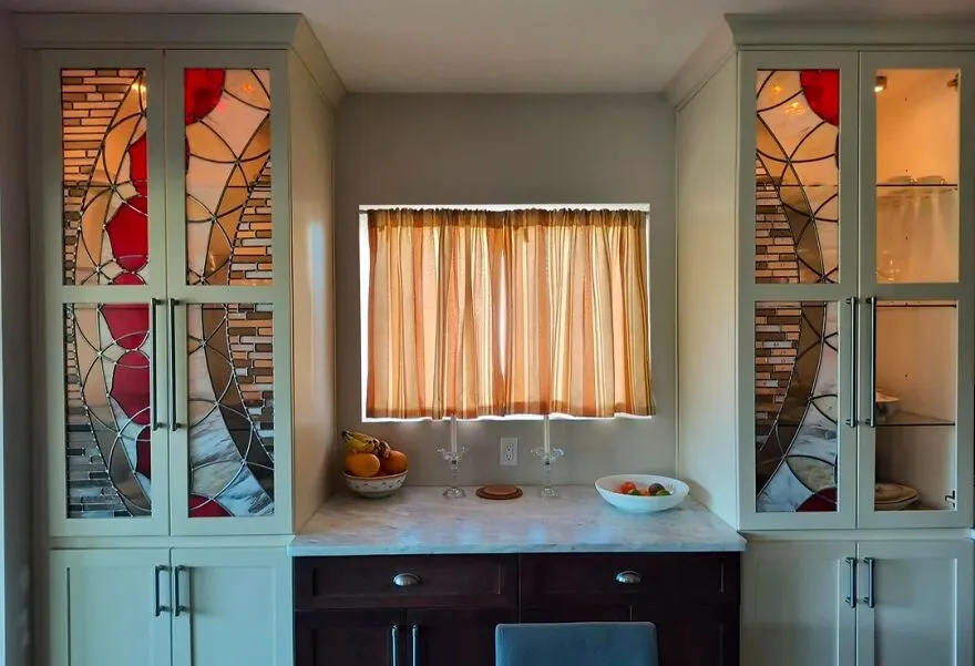 Stained glass kitchen cabinets with colorful patterns flank a window, showcasing modern kitchen cabinet design inspiration.