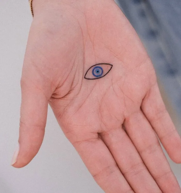 Small hand tattoo featuring a blue eye design, centered on an open palm.