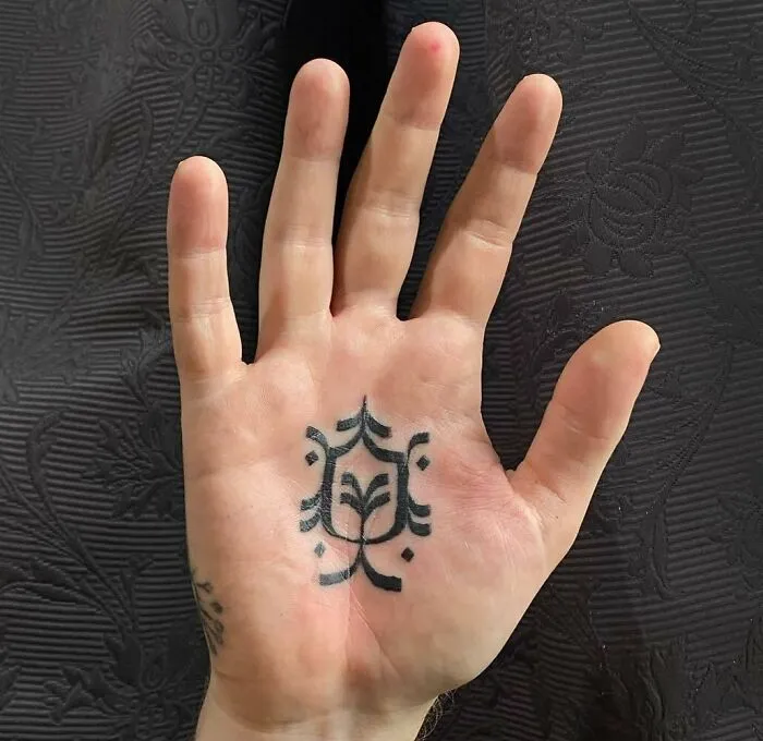 A small hand tattoo featuring a simple geometric pattern on the palm against a textured black background.