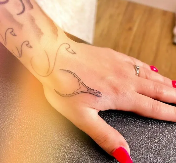 Small hand tattoo of a wishbone on the back of a hand with red nails and a silver ring, showcasing delicate design.