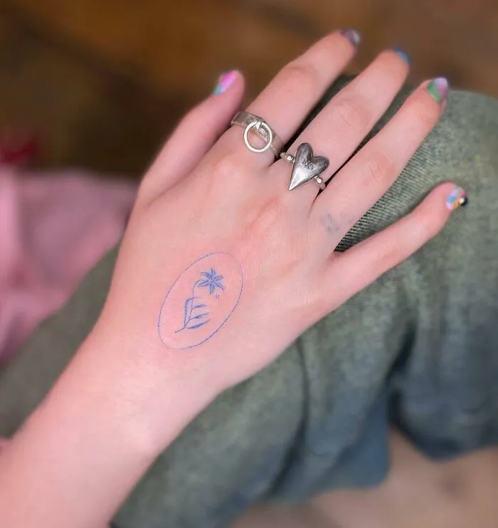 Small hand tattoo featuring a delicate flower in blue ink with silver rings, highlighting creative design on hand.