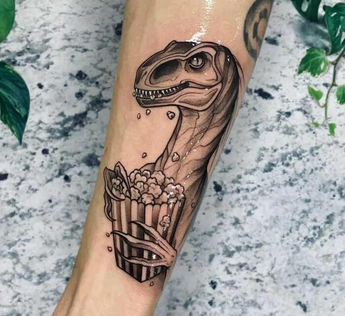 Dinosaur tattoo featuring a velociraptor holding popcorn, expertly inked on an arm with a textured background.