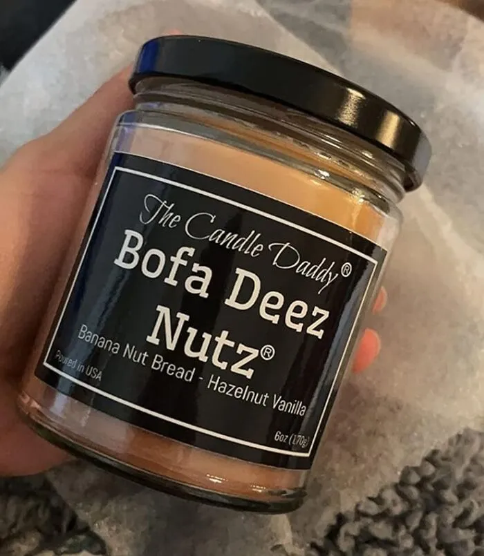 Hand holding a humorous scented candle labeled "Bofa Deez Nutz," featuring banana nut bread and hazelnut vanilla scent.