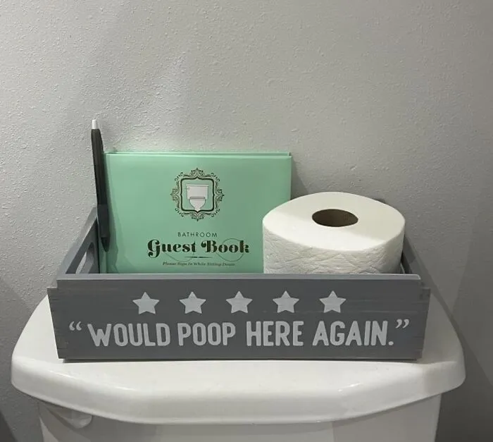 Bathroom guestbook and toilet paper with humorous sign, "Would poop here again," in a quirky bathroom setting.