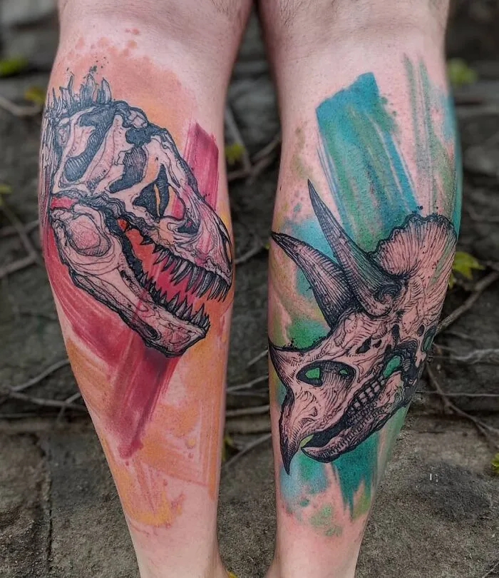 Colorful dinosaur tattoos on legs featuring skull designs of a T. rex and a triceratops.