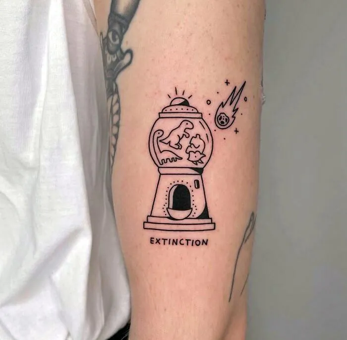 Dinosaur tattoo featuring a gumball machine with dinosaurs and meteors, labeled "EXTINCTION," on a person's arm.
