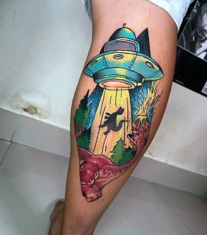 Vibrant dinosaur tattoo featuring UFO abduction theme on a person's leg.