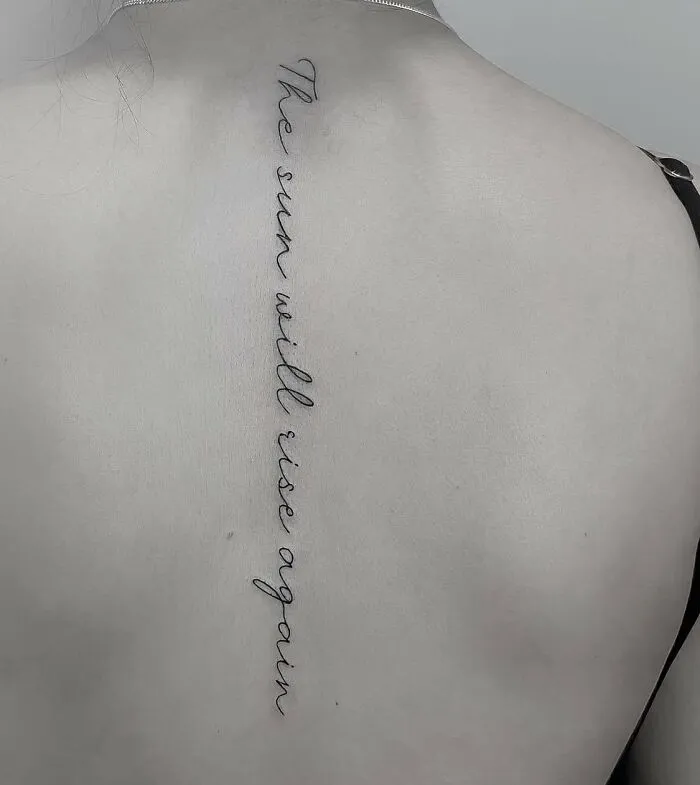Elegant script spine tattoo with the phrase "The sun will rise again."