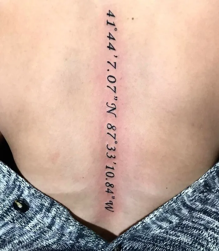 Spine tattoo featuring geographic coordinates in black ink on skin.