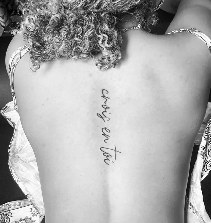 Curly-haired person with a beautiful spine tattoo reading "crois en toi" in elegant script.
