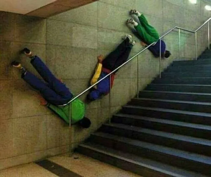 People in colorful outfits lying sideways on a wall alongside a staircase, creating a bizarre and cursed visual effect.
