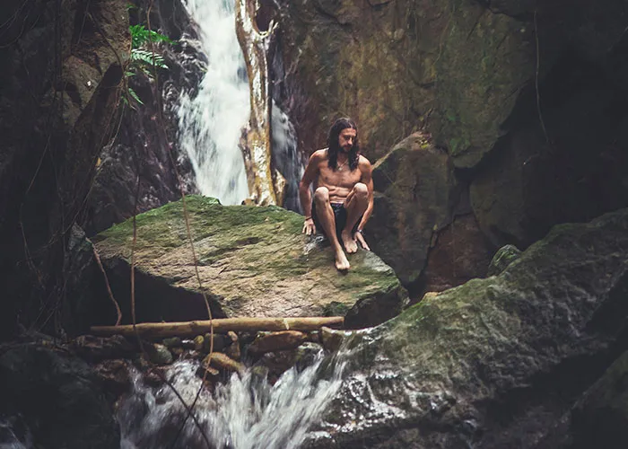 A person sitting on a rock near a waterfall, surrounded by nature, creating an amusing scene despite the serene setting.