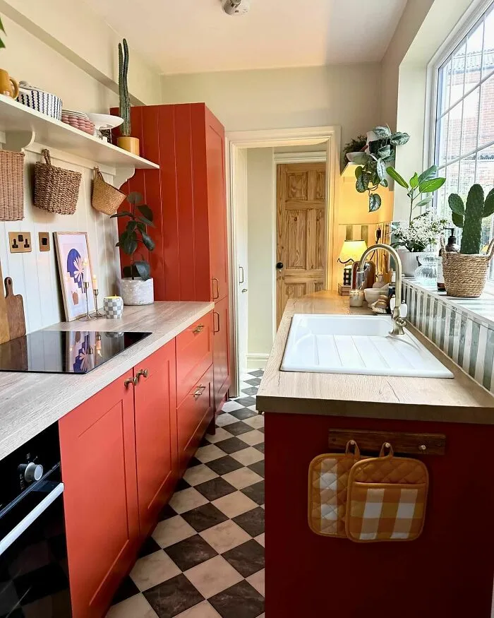 Bright galley kitchen with red cabinets, checkered floor, wooden countertops, and natural light from large window.