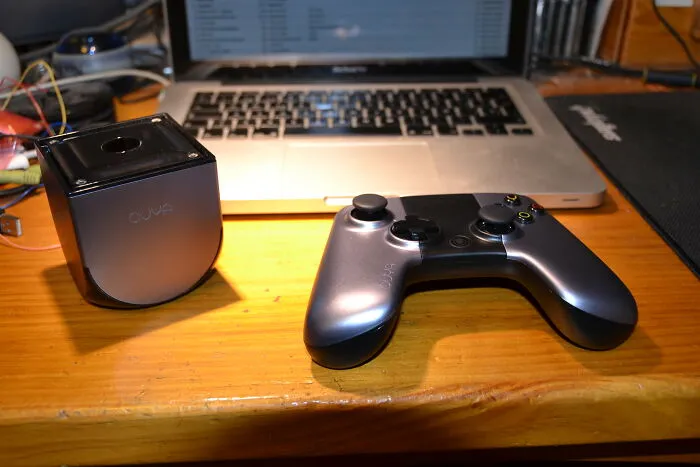 A failed gaming console and controller on a wooden desk with a laptop in the background.