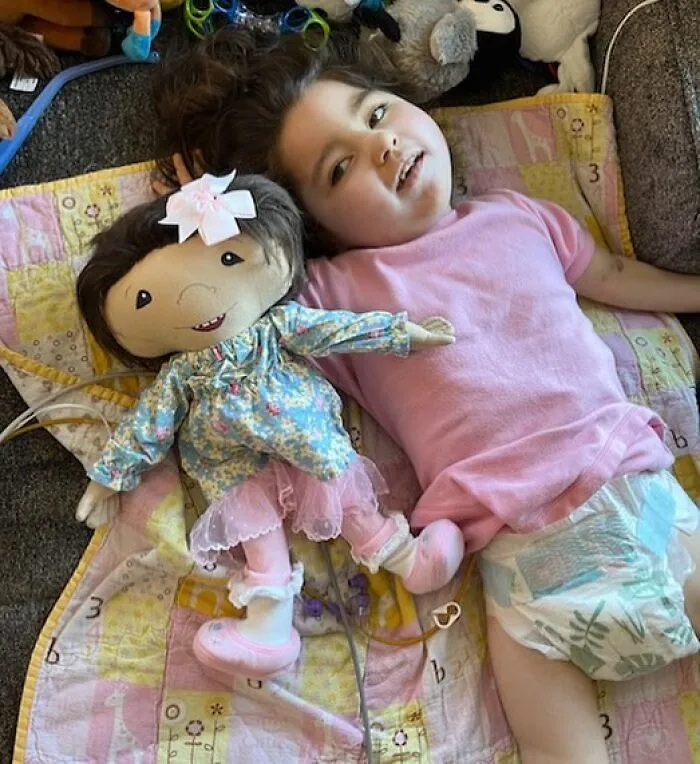 Inclusive dolls: A young girl with rare health conditions lies next to her custom doll, both with feeding tubes.