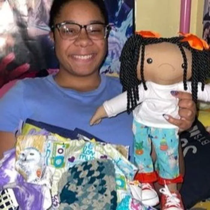 A smiling woman wearing glasses holds an inclusive doll with braided hair and colorful clothes, representing rare health conditions.