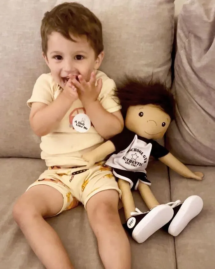 A smiling boy with dark hair sits next to an inclusive doll with matching hair and outfit, representing rare health conditions.