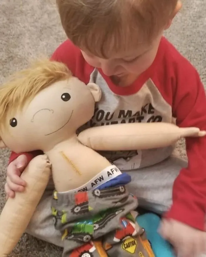 A young boy looking down at his inclusive doll, which has a medical tube on its stomach, representing rare health conditions.
