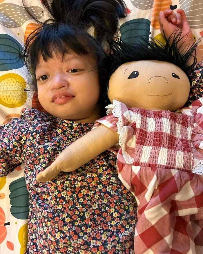 A girl with health conditions smiling next to her inclusive doll that represents her.
