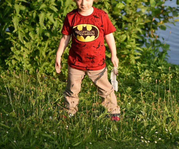 A child outdoors wearing a playful red "Ratman" shirt, standing on grass with greenery in the background.