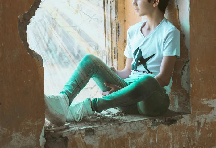 Teen sitting in a damaged window frame, wearing a printed shirt and jeans, symbolizing unfortunate names theme.