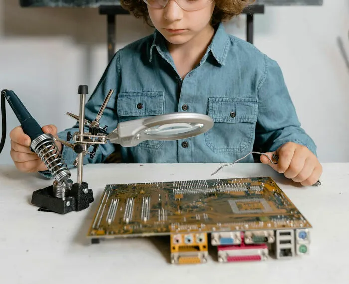 Child in glasses and denim shirt soldering a circuit board, representing creativity and early STEM education.