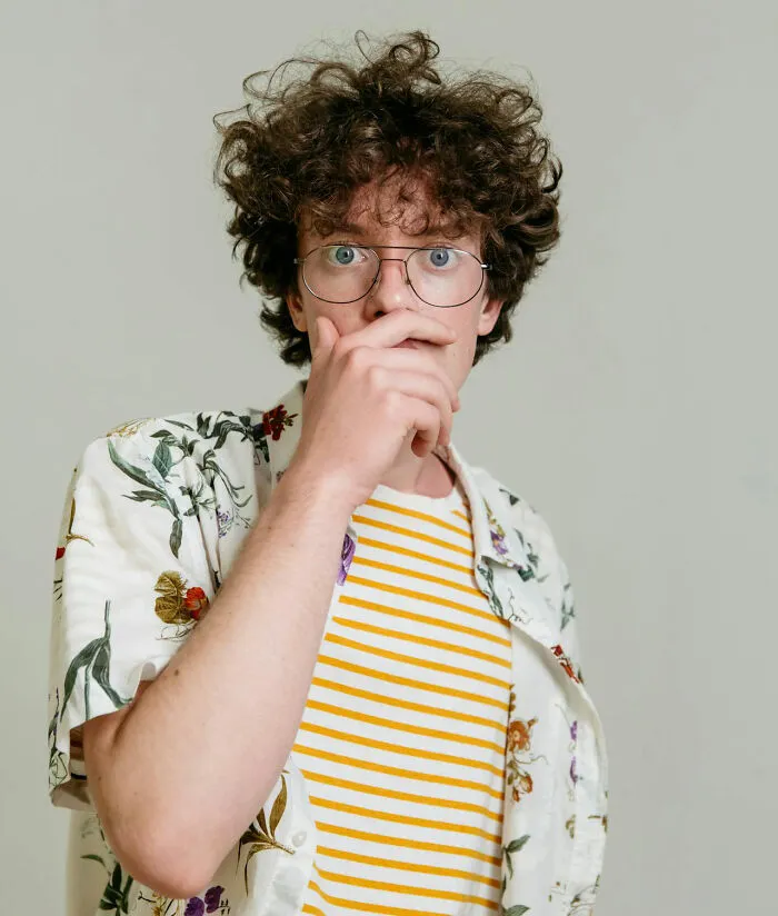 Young man in glasses wearing floral shirt, hand covering mouth, reacting humorously to unfortunate baby names.