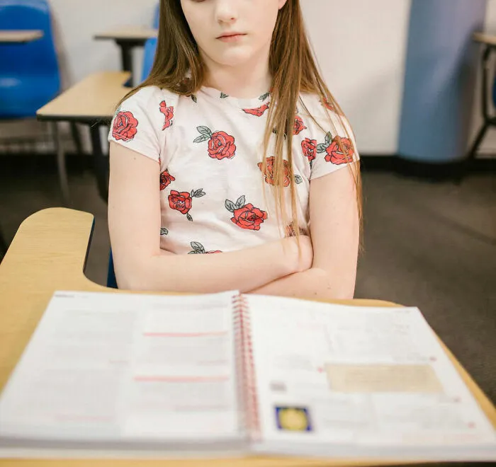 Young girl in classroom, arms crossed, notebook open in front, reflecting on hilariously unfortunate baby names.