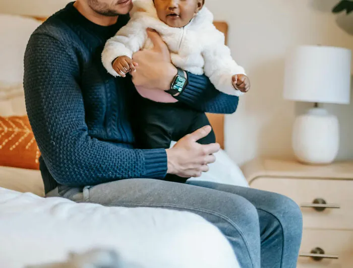 A man holding a baby on his lap in a cozy bedroom, wearing casual knitwear, with a lamp and dresser in the background.