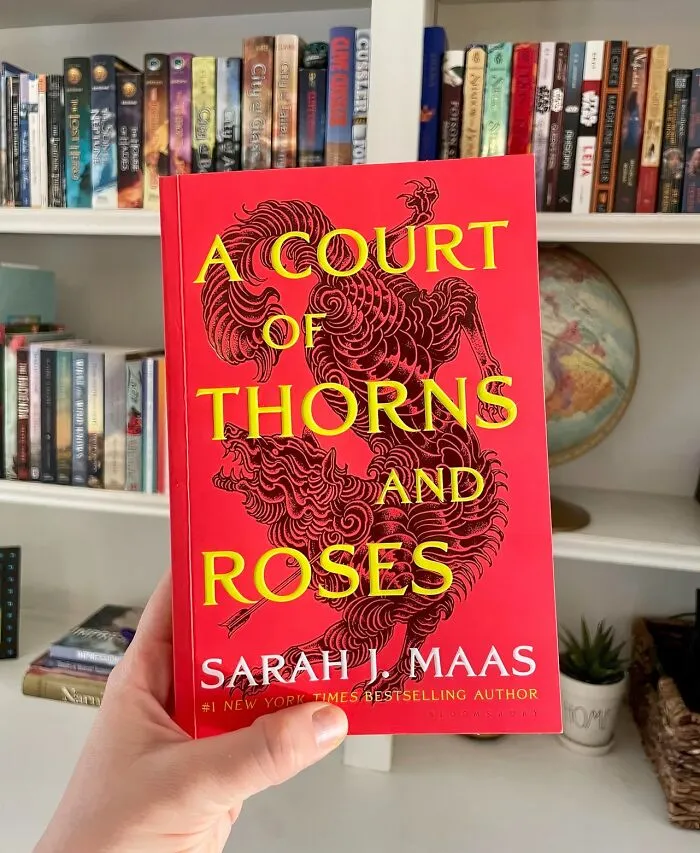 Red cover of "A Court of Thorns and Roses" held in front of a bookshelf, a perfect cheerful gift idea.