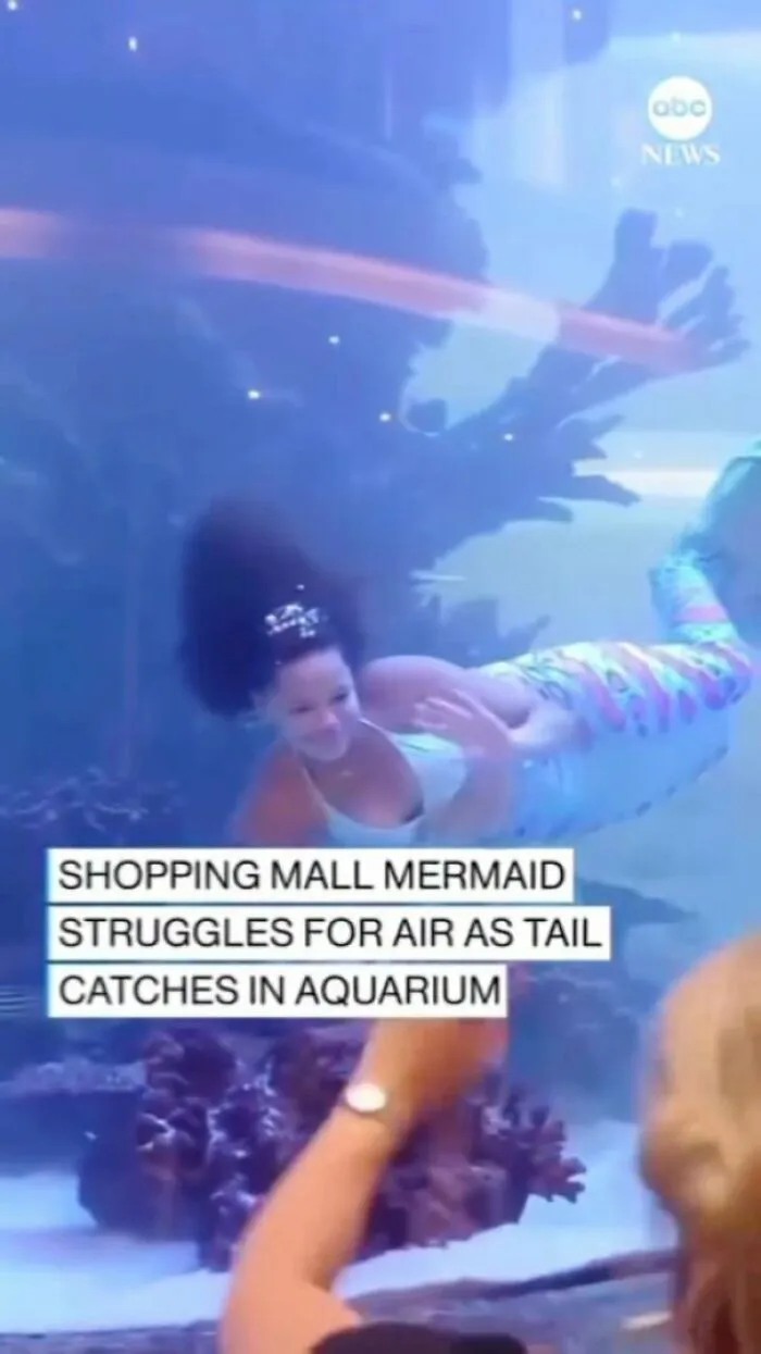 Mermaid performer struggles in aquarium tank at shopping center, showcasing challenges of capitalism.