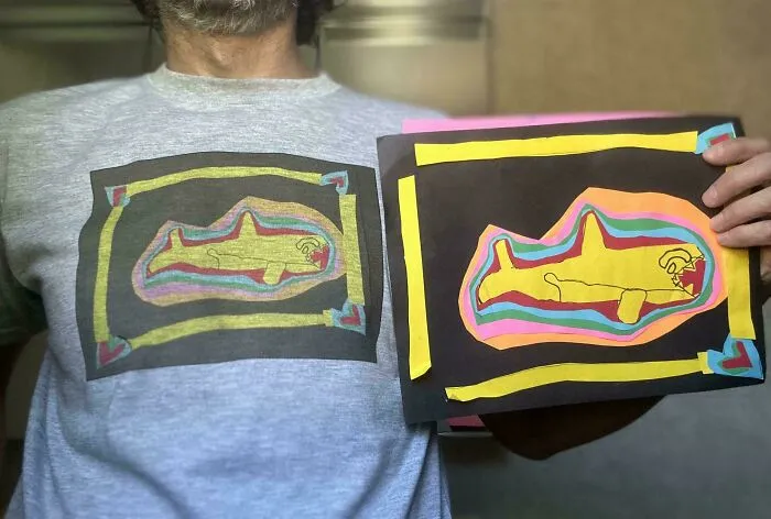 Man wearing a gray shirt with a colorful shark drawing matching the handmade shark artwork he is holding, showcasing funny dad moments.
