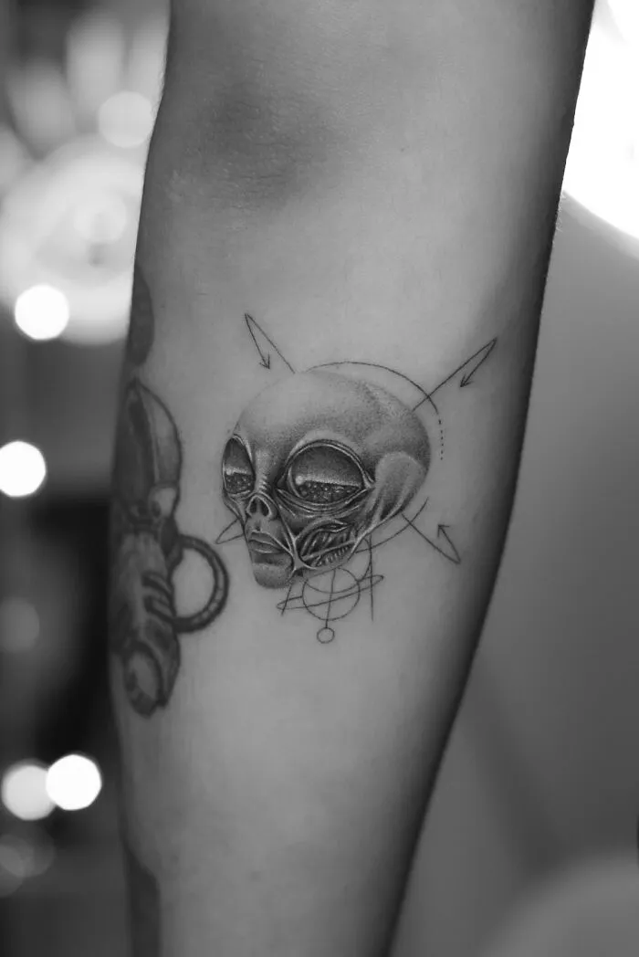 Single needle tattoo of an alien with geometric patterns on forearm, showcasing intricate design and shading.