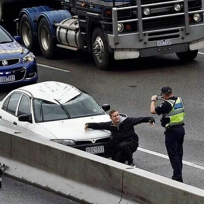 Hombre posando de forma incómoda frente a un policía y un coche blanco en una carretera transitada.