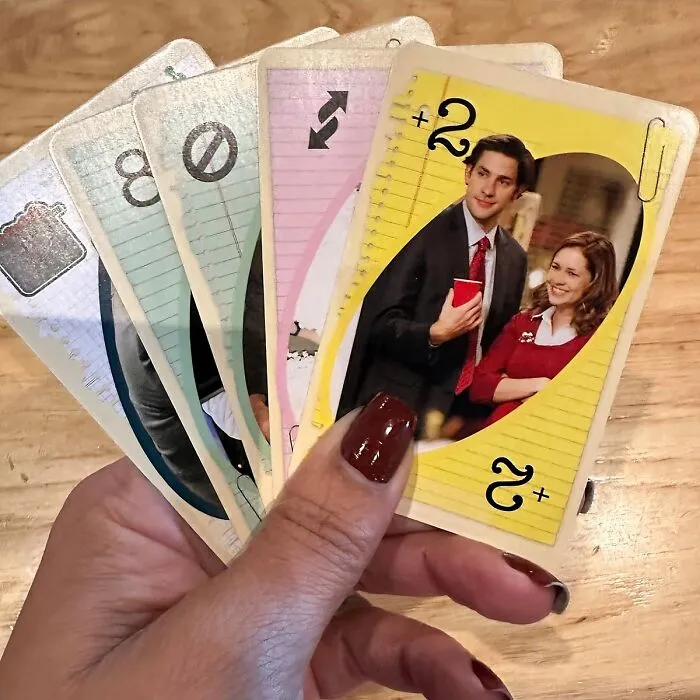 Hand holding work supplies-themed UNO cards with office characters, on a wooden table.