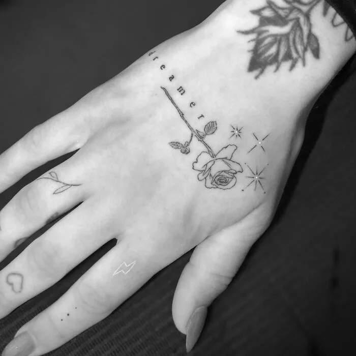 Hand with single needle tattoos: rose, "dreamer" text, and small symbols on fingers.