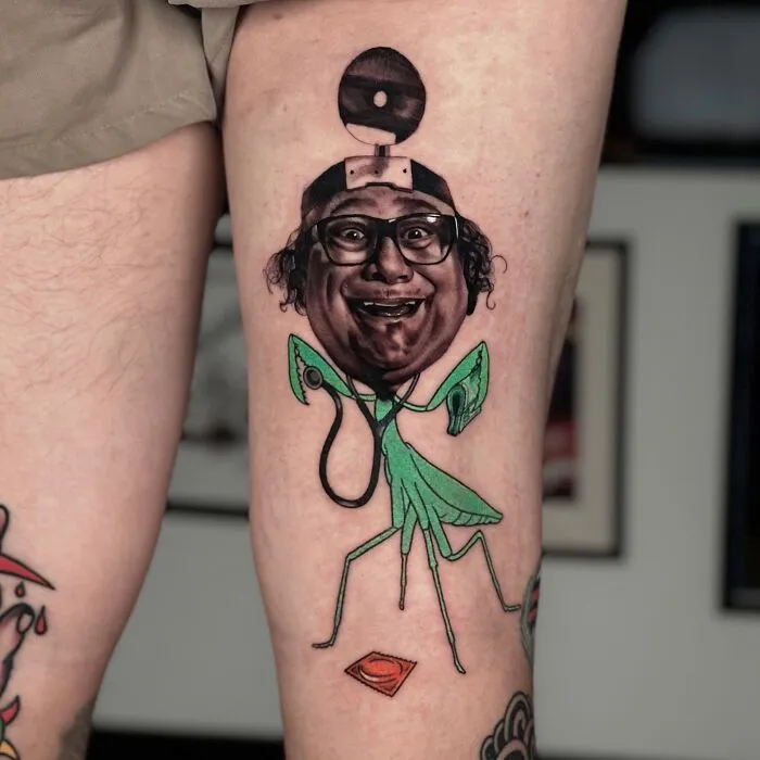 Tattoo merging two artistic styles, featuring a humorous portrait with a mantis body, on a person's thigh.