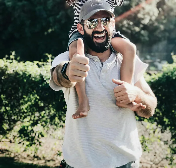Man wearing sunglasses and cap giving a thumbs up, carrying child on shoulders, epitomizing dad humor outdoors.
