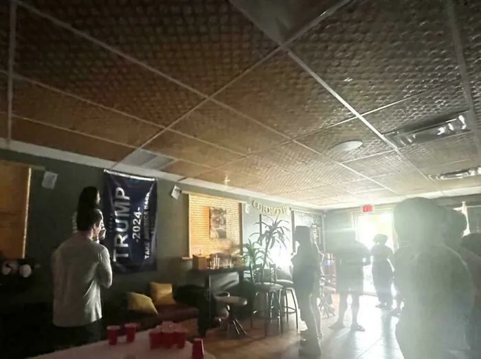 People in a dimly lit room with a political banner and cups on a table, evoking a boring dystopia theme.