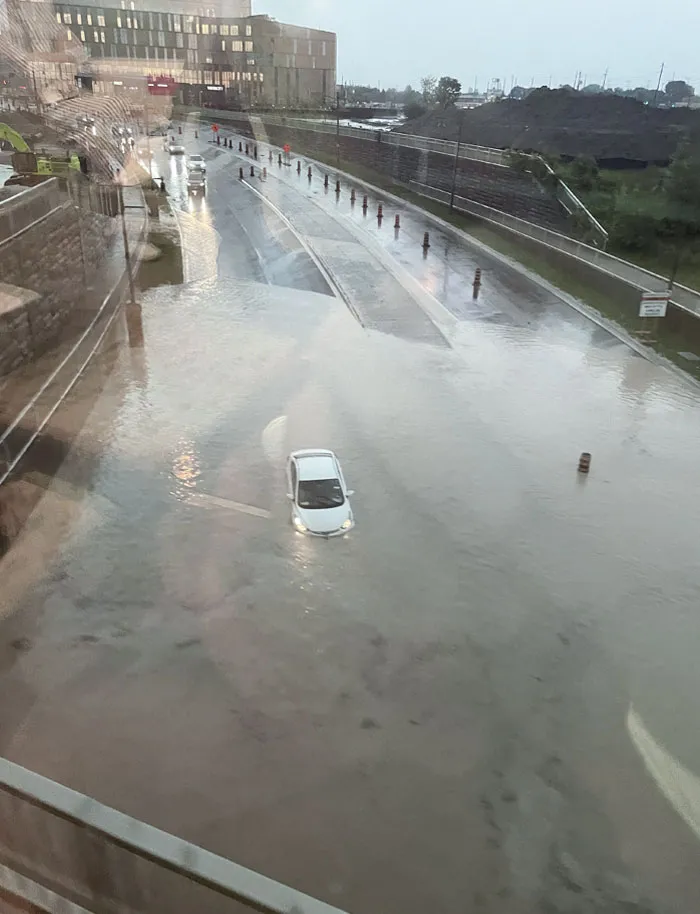 Flooded urban street with a white car partially submerged under rising water during heavy rainstorm.