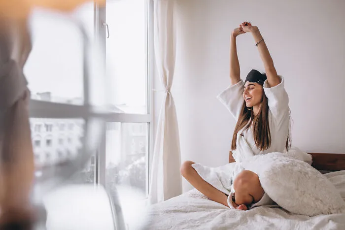 Young woman stretching happily on bed in morning light, enjoying simple things people started appreciating after getting older.