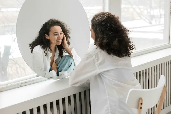 Woman with curly hair appreciating herself in a mirror by a window, reflecting self-love and aging appreciation.