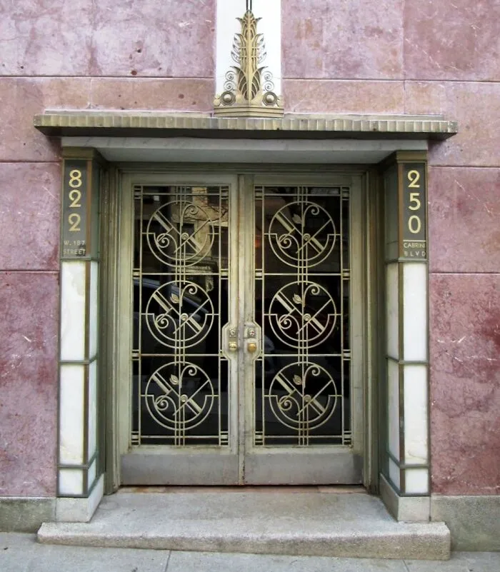 Intricate Art Deco doors with geometric metalwork and gold accents framed by marble and stone at a historic building entrance.
