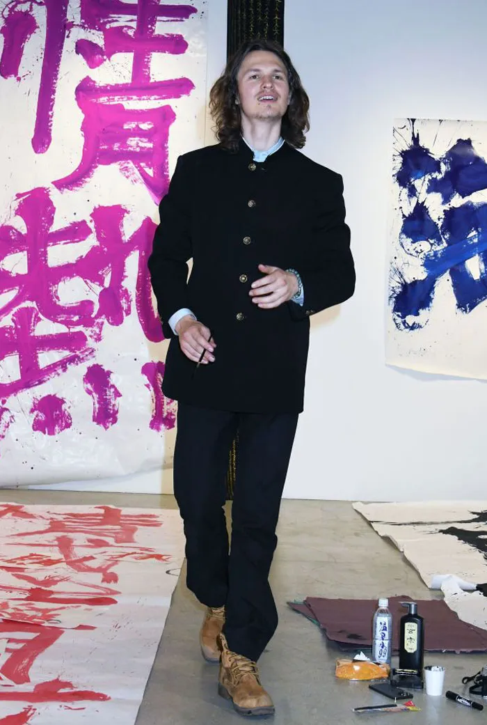  A B list celebrity, Ansel Elgort, is standing in an art gallery, wearing a black button-up jacket and boots, with large abstract paintings in the background. He is holding a paintbrush during the event.