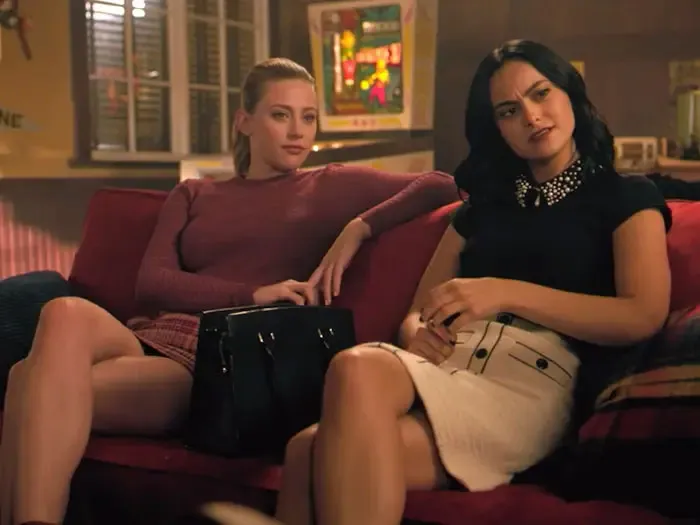 Two women sitting on a red couch, representing iconic duos, in a cozy room setting.