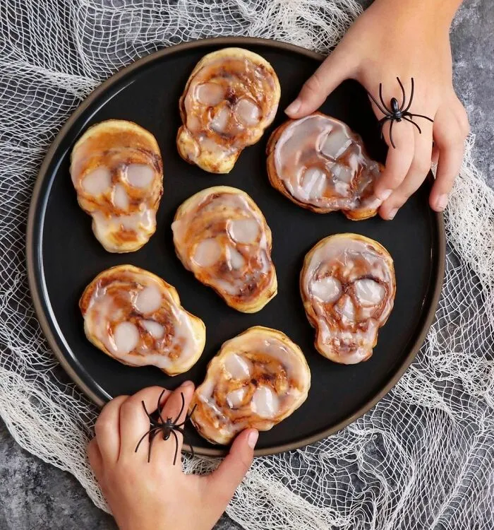 Skull-shaped Halloween snacks on a black plate with children’s hands wearing spider rings reaching for them.