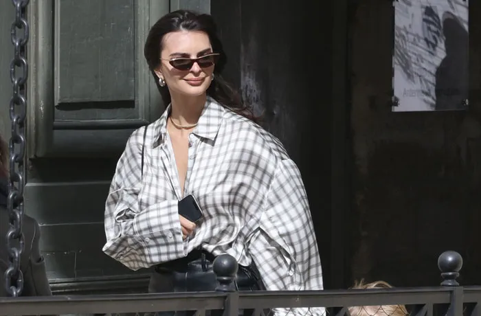  A B list celebrity, Emily Ratajkowski, is walking outdoors wearing sunglasses, an oversized plaid shirt, and holding a phone, with a subtle smile.