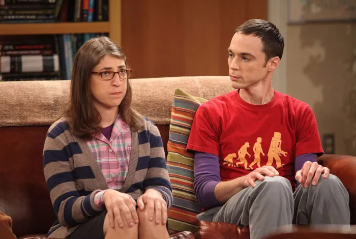 Two characters sitting on a couch, one wearing glasses and a striped sweater, the other in a red graphic tee. Iconic duos moment.