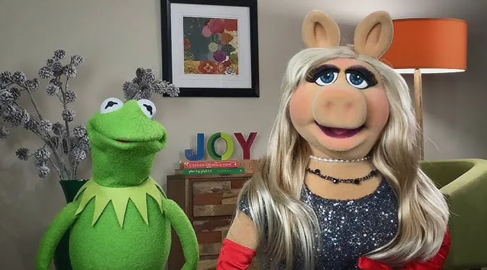 Iconic puppet duo posing in a cozy living room, decorated with artwork and a lamp in the background.